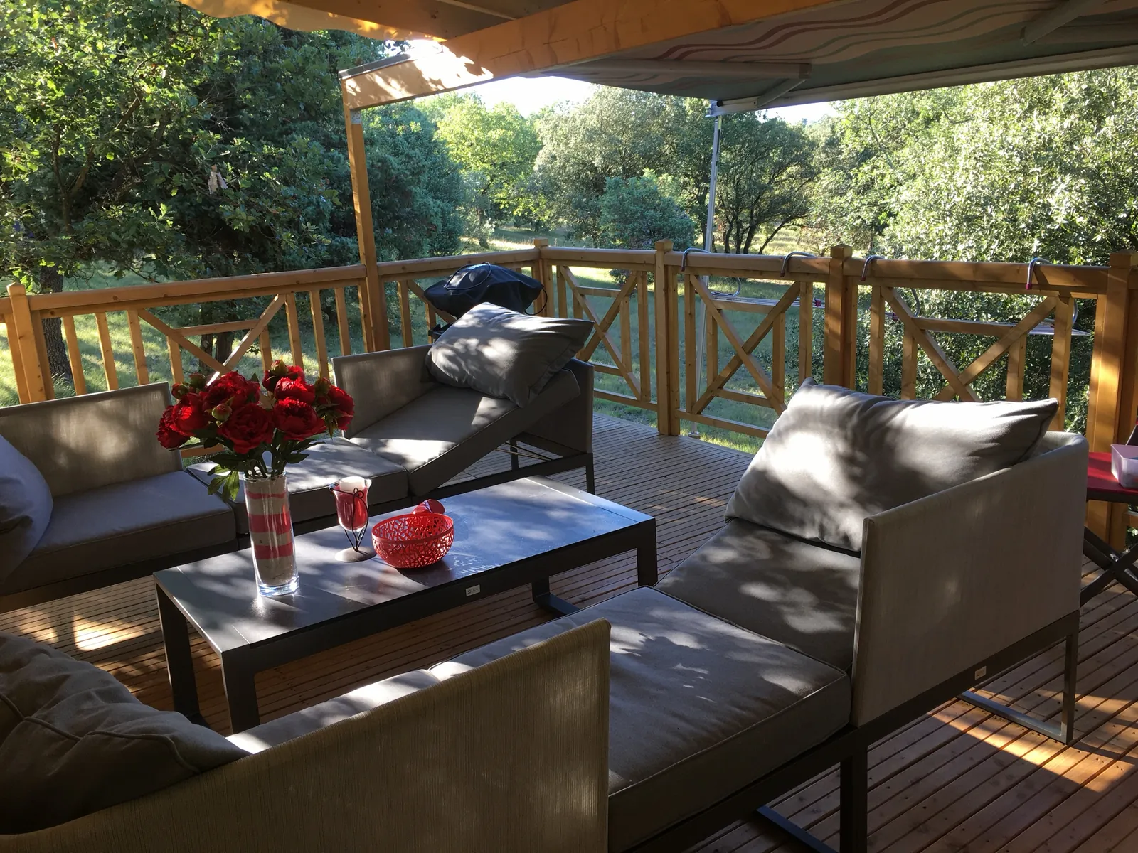 Salon d'extérieur installé sur une terrasse couverte en bois, sous les arbres, avec garde-corps à croix de Saint-André