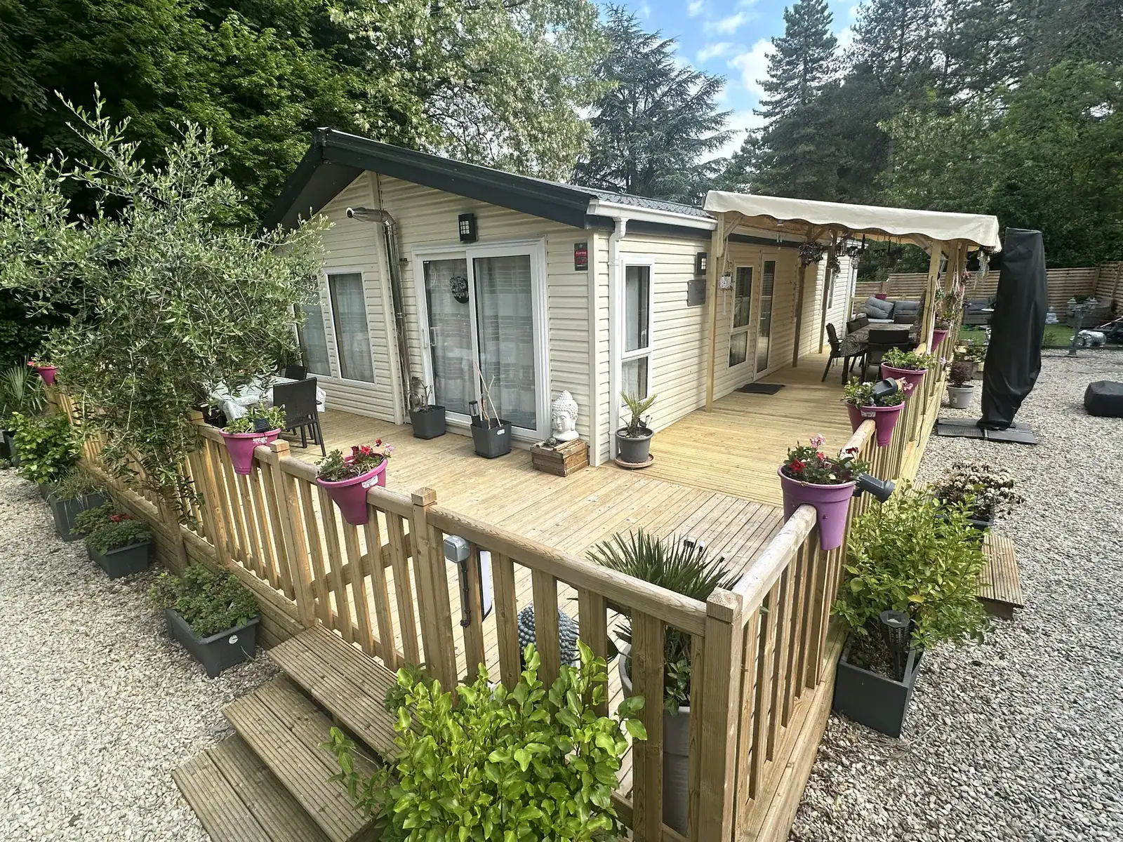 Terrasse en bois en forme de L entourant deux côtés d'un mobil-home crème, avec garde-corps barreaudé et jardinières