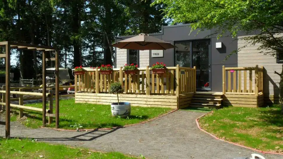 Terrasse simple en bois devant un mobil-home contemporain gris, avec garde-corps barreaudé, jardinières de géraniums et marches d'accès