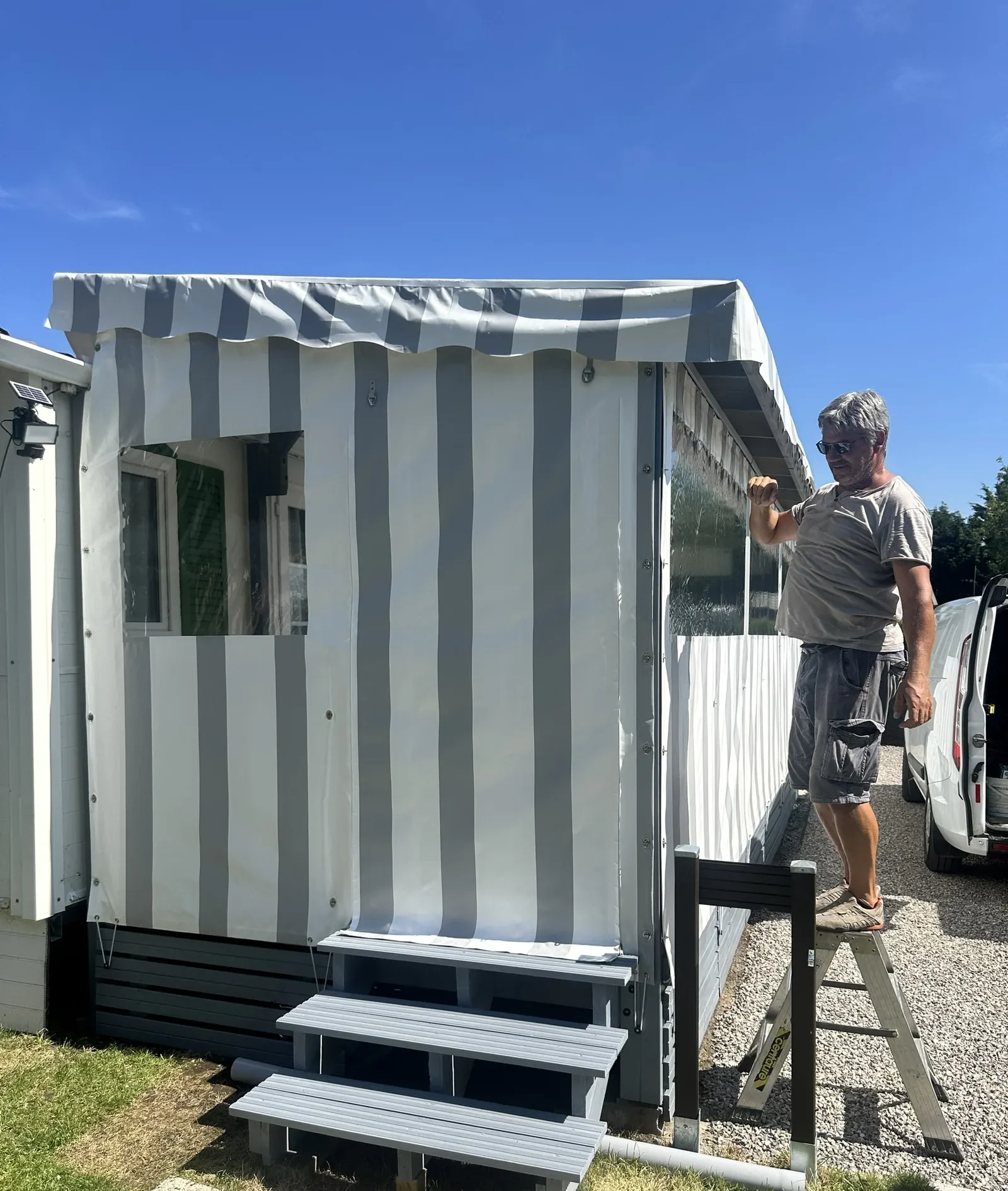 Poseur ajustant depuis un escabeau la bâche rayée grise d'une terrasse couverte, pendant l'installation