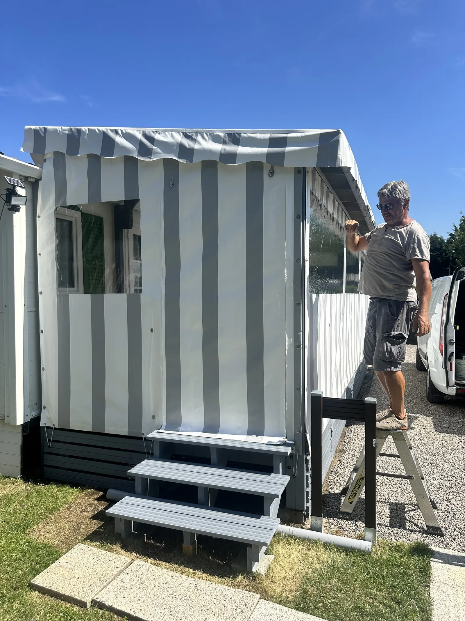 Poseur ajustant depuis un escabeau la bâche rayée grise d'une terrasse couverte, pendant l'installation
